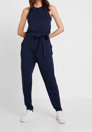 Kobieta w granatowym, bezrękawnym kombinezonie z paskiem przewiązanym w talii i białych platformowych trampkach, stojąca na tle jednolitej białej ściany.