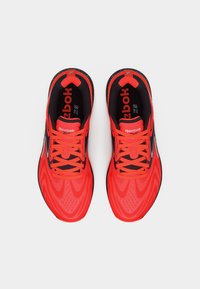 Ζευγάρι λαμπερά κόκκινα και μαύρα παπούτσια τρεξίματος Reebok με κορδόνια, σε θέα από πάνω σε λευκό φόντο.