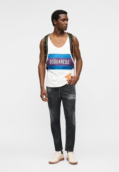 Homme portant un débardeur blanc Dsquared2, un jean foncé, des baskets blanches et un sac à dos vert, debout devant un fond uni.