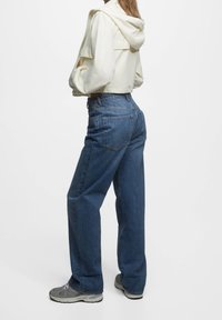 Sweat à capuche court beige clair avec une capuche, associé à un jean denim bleu ample et des baskets grises. Le jean présente des poches arrière et une silhouette décontractée.