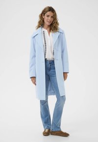 Cappotto di lana azzurro chiaro con colletto oversize, davanti doppiopetto, abbinato a una blusa bianca e jeans flare in denim. Sneakers marroni.