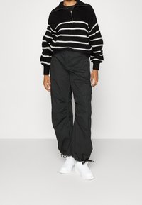 Schwarz-weißer gestreifter Strickpullover mit einem halben Reißverschlusskragen, kombiniert mit lockeren schwarzen Cargo-Hosen und weißen Sneakers.