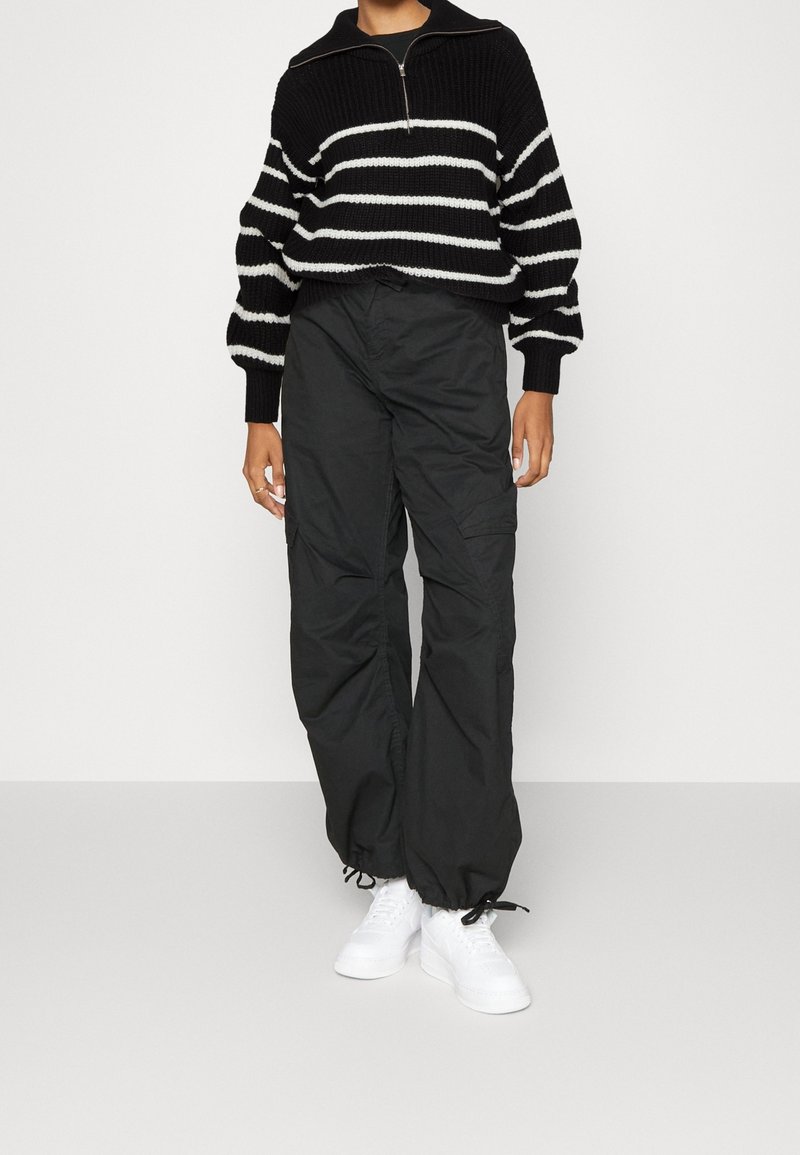 Schwarz-weißer gestreifter Strickpullover mit einem halben Reißverschlusskragen, kombiniert mit lockeren schwarzen Cargo-Hosen und weißen Sneakers.