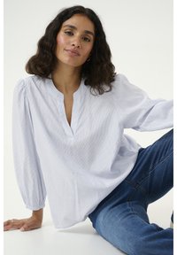 Blusa a righe blu chiaro con scollatura a V, maniche a sbuffo e sottili punti testurizzati. Indossata con jeans blu, posata su uno sfondo chiaro.