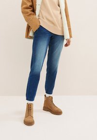 Personne portant un jean bleu, des bottes à lacets couleur bronzée, un sweat-shirt beige et une veste doublée de peau retournée marron, debout sur un sol blanc contre un mur uni.