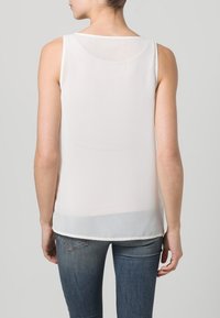 Blusa bianca senza maniche con scollatura a barchetta, tessuto trasparente e orlo dritto, abbinata a jeans aderenti.