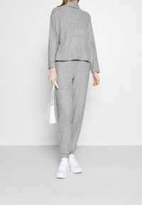 Jasnoszara, długofalowa bluzka z wysokim kołnierzem i dopasowanymi spodniami z troczkiem. Miękka, teksturowana tkanina; w zestawie z białymi sneakersami i małą torbą.