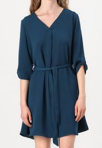 Robe bleu sarcelle en tissu doux, dotée d'un col en V, de manches trois-quarts avec revers roulés, et d'une ceinture en tissu à la taille.