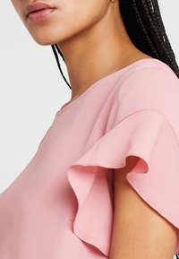 Haut rose à manches courtes avec des accents froncés le long des manches, en tissu lisse, avec un col rond, doté d'un design léger.