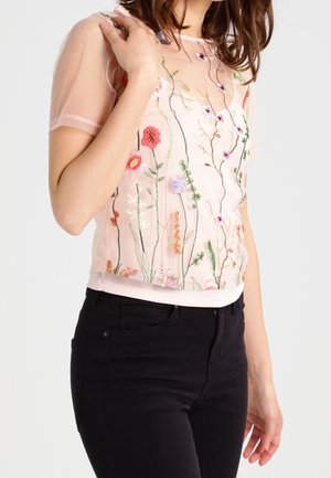 Vrouw draagt een doorschijnende roze top met kleurrijke geborduurde bloemen en zwarte broek met hoge taille, poseert met één hand bij haar schouder.
