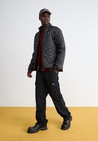 Melnā siltinātā jaka ar stāvošu apkakli, apvienota ar tumši sarkanu pogājamo kreklu un melniem cargo biksēm. Modelis valkā melnas platformas kurpes.