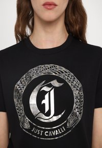 T-shirt noir en coton avec un design de serpent métallique argenté et un logo comprenant le texte "JUST CAVALLI" disposé en cercle.