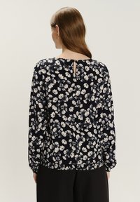 Frau mit langen braunen Haaren, die eine schwarze Bluse mit weißen Blumen und schwarze Hosen trägt, von hinten vor einem schlichten Hintergrund gesehen.