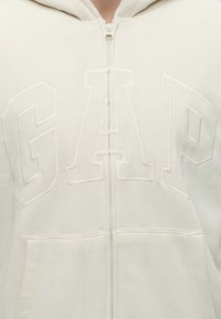 Sweat à capuche crème zippé avec des lettres brodées en relief formant GAP sur la poitrine et une poche kangourou à l'avant.