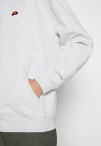 Sudadera gris claro con un pequeño logotipo bordado multicolor, bolsillos laterales, puños y dobladillo de canalé; textura de tela suave.