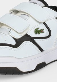 Λευκά παιδικά αθλητικά παπούτσια με μαύρες λεπτομέρειες, δύο λουράκια με βέλκρο και πράσινο λογότυπο κροκόδειλου στο πλάι.