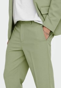 Pantalones ajustados verde claro de tela suave, con bolsillos laterales y un ajuste de altura media, complementados con una camisa blanca lisa.