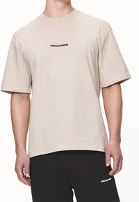 Mies, joka pitää yhen beigeä lyhythihaista t-paitaa, jossa on "PEGADOR"-teksti rinnassa, ja mustia housuja, joissa on "PEGADOR"-teksti vasemmassa reidessä.