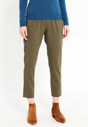 Persoon met olijfgroene cropped broek, een blauw lang mouw shirt en bruine suède enkellaarsjes staand op een witte achtergrond.