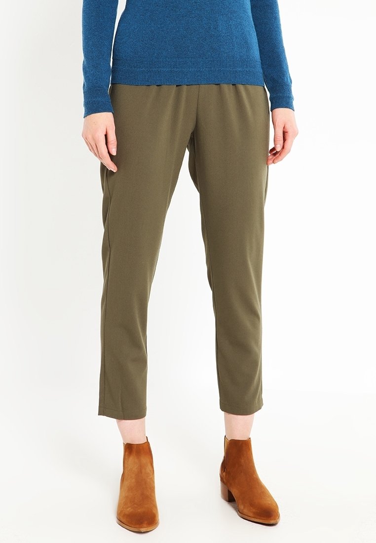 Personne portant un pantalon court vert olive, un haut bleu à manches longues et des bottines en daim marron, debout sur un fond blanc.