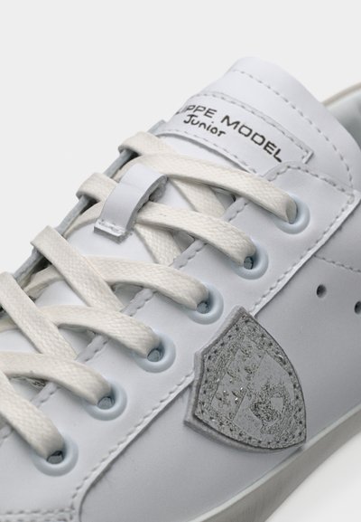 Baskets blanches avec des lacets blancs, une étiquette "Pepe Model Junior" sur la languette, et un écusson argenté brodé sur le côté.