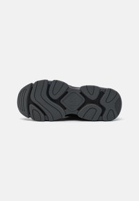 Black athletic shoe outsole featuring a thick tread pattern with varied textures, grooves, and a central logo detail.