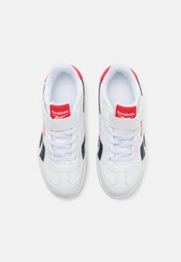 Zapatillas blancas con un acabado de cuero suave y detalles en azul marino. Presentan un logotipo rojo y una correa de velcro además de los cordones tradicionales.