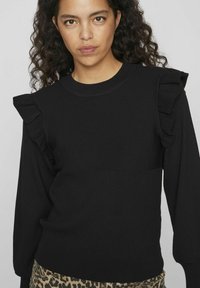 Melna džemperis ar ribinātu tekstūru, izdaiļots ar drapējumiem uz pleciem un garām piedurknēm. Ikdienišķi valkāts virs rakstainām leopardu-print biksēm.