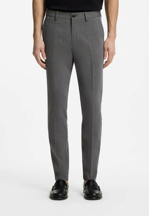 Mężczyzna w obcisłych szarych spodniach i czarnych mokasynach, stojący na białym tle.