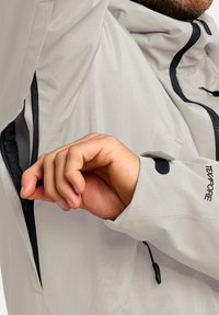 Giacca impermeabile grigio chiaro con tasca con zip sotto il braccio, polsini regolabili e dettagli neri sulla zip e sul logo.