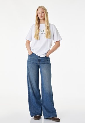 Joven mujer con el pelo largo y rubio que lleva una camiseta blanca con estampado floral, vaqueros azules de pierna ancha y zapatos marrón oscuro, con las manos en los bolsillos.