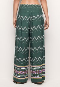 Green wide-leg pants featuring a zigzag white pattern, smocked waist, and colorful geometric accents at the hem. Lightweight fabric, relaxed fit.