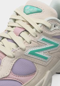 Κοντινό πλάνο μπεζ και λιλά αθλητικού παπουτσιού με κρεμ κορδόνια και τιρκουάζ λογότυπο "N", που δείχνει λεπτομέρειες υφής από δίχτυ και καστόρι.