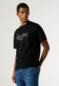 Černé bavlněné tričko s krátkým rukávem a kulatým výstřihem. Na hrudi je bílé, opotřebované logo. Komplet doplněné modrými džíny.