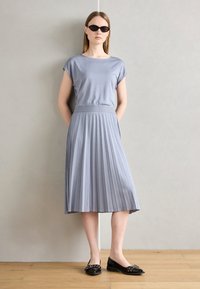 Szürke, redőzött ruha rövid ujjal, sima anyagból, és kényelmes derékkal. Fekete, zárt orrú balerinacipővel és elegáns fekete napszemüveggel párosítva.