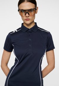 Mörkblå polo skjorta med vita detaljer, korta ärmar och krage. Har en knäppning framtill och en slimmad, atletisk design. Åtföljd av skyddsglasögon.