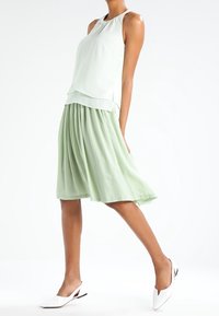 Světle zelená, bezramenná blůzka s kulatým výstřihem, spárovaná s volnou, plisovanou sukní. Bílé baleríny dokončují outfit. Hladká textura látky.