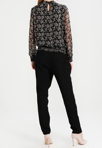 Blouse noire à fleurs avec des manches longues transparentes, détail en forme de goutte dans le dos, associée à un pantalon noir ajusté et des chaussures à talons nude.