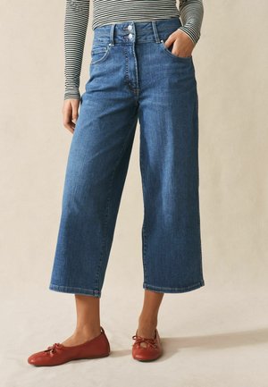 Frau trägt hoch taillierte, verkürzte, weitbeinige blaue Jeans, gestreiftes Langarmshirt und rote Ballettschuhe, steht mit einer Hand in der Tasche.