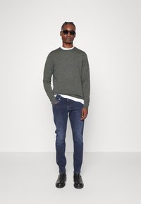 Maglione grigio a maglia con scollo a girocollo, abbinato a jeans slim-fit blu scuro. Il modello indossa occhiali da sole neri e stivali neri alla caviglia.