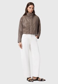 Ruskea nahkatakki, jossa korkea kaulus, yhdistettynä valkoisiin leveälahkeisiin housuihin ja ruskeisiin kuvioituihin balleriinoihin. Sileä pinta, minimaalinen metalliosat.