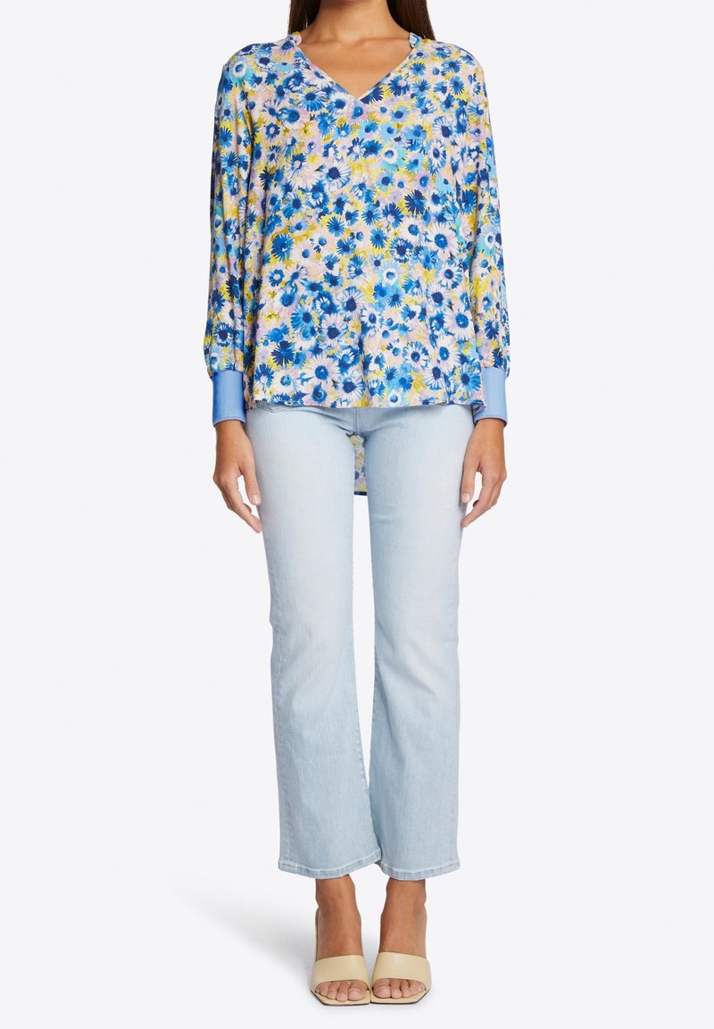 Blouse fleurie à manches longues en bleu, rose et jaune avec un col en V et des poignets bleus, associée à un jean évasé bleu clair et des sandales beiges.