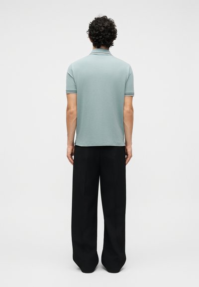 Άνδρας με σγουρά σκούρα μαλλάκια που φοράει ανοιχτό πράσινο πόλο με κοντά μανίκια και φαρδιά μαύρη παντελόνια, στέκεται με την πλάτη προς τα εμπρός σε απλό φόντο.