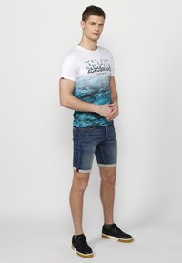 Short-sleeve white T-shirt with blue gradient design and text, paired with denim shorts featuring rolled cuffs and patterned accents, worn with black sneakers.