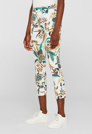 Dopasowane, krótkie spodnie z kolorowymi wzorami kwiatów i liści, noszone z białymi trampkami, na stojącej osobie.