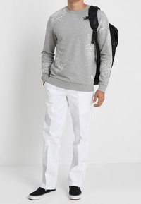 Felpa grigia con disegni floreali bianchi, abbinata a pantaloni bianchi e scarpe da ginnastica nere. La modella tiene uno zaino nero.