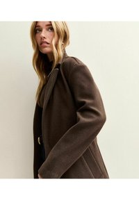 Abrigo de lana marrón con un diseño ajustado, que presenta solapas en pico, bolsillos laterales y una textura suave, usado sobre una parte superior oscura.