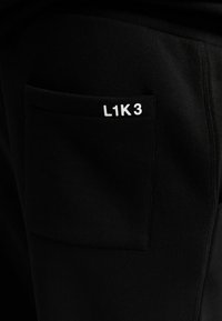 Black fabric joggers featuring a square pocket with the text “L1K3” in white. The material appears soft with a smooth texture.