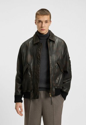 Brown leather bomber jacket with a smooth texture, featuring a collar, front pockets, and ribbed cuffs. Worn over a dark sweater.