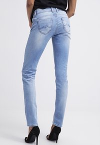 Světle modré džíny s úzkým střihem, vybledlou texturou a dvěma zadními kapsami s zakřivenými švy. Nosené s černými vysokými podpatky.